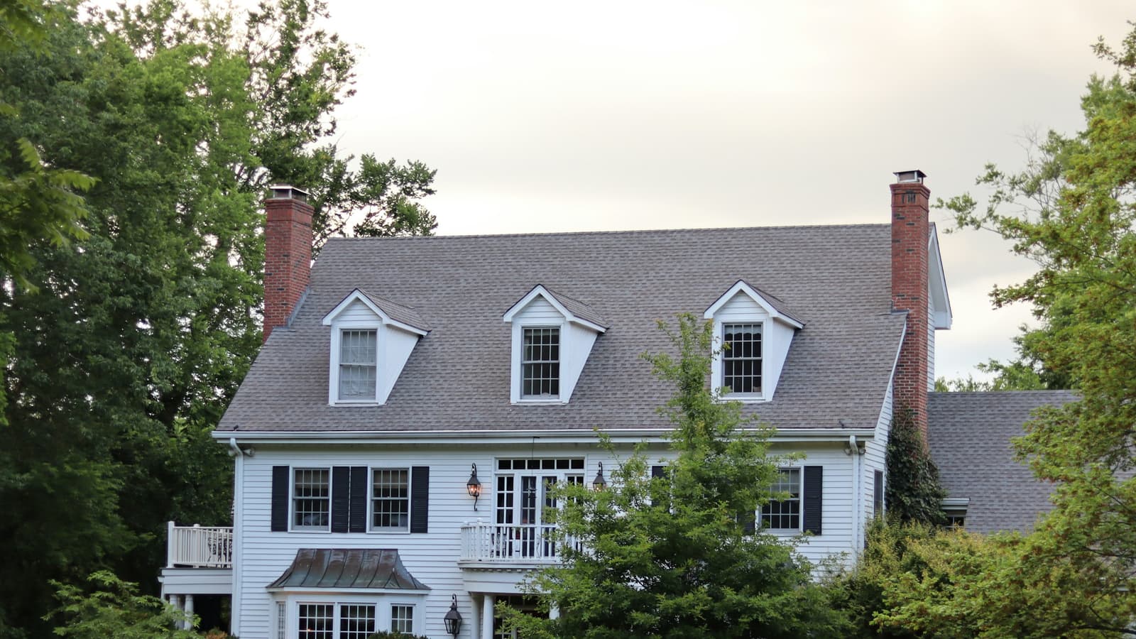 White two-story colonial home with a gray asphalt-shingle roof, dormers, brick chimneys and a green lawn framed by leafy trees